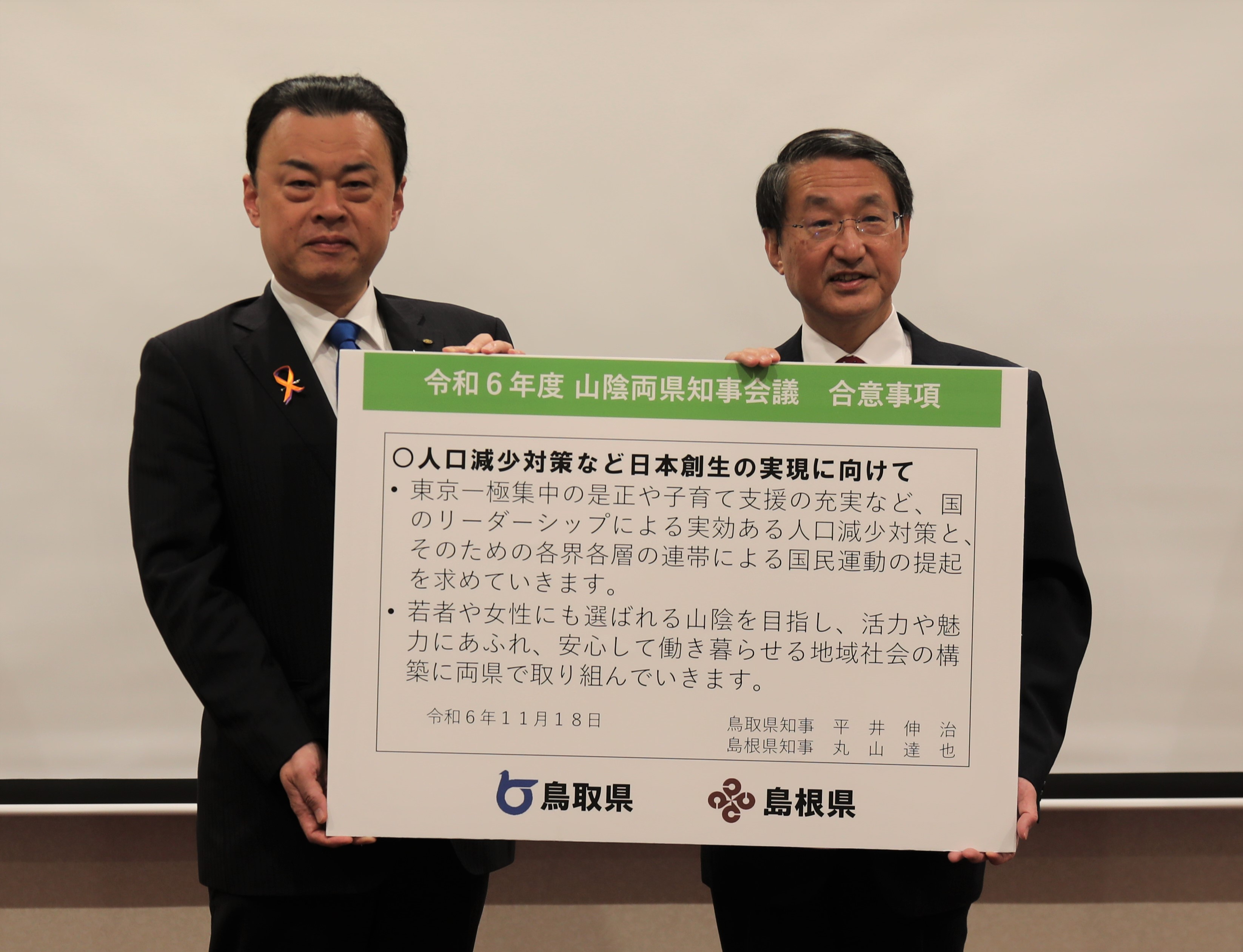 合意事項が印刷されたパネルを掲げる両県知事